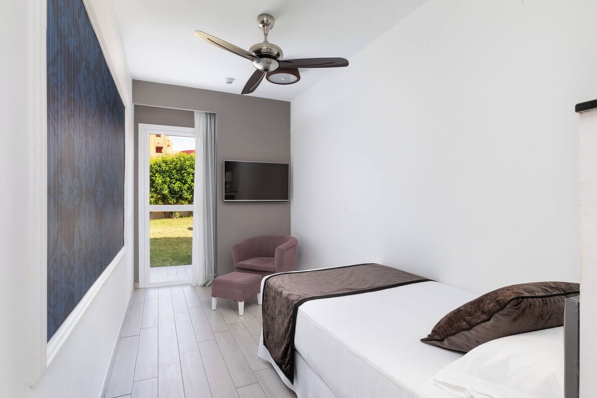 A modern bedroom with a bed, a ceiling fan, a TV mounted on the wall, and a view of the outdoors.