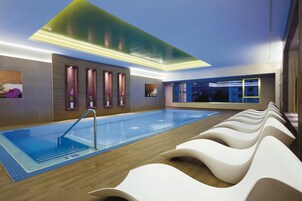 A modern indoor swimming pool with a wooden floor, a glass barrier, and a row of white chairs along the side.