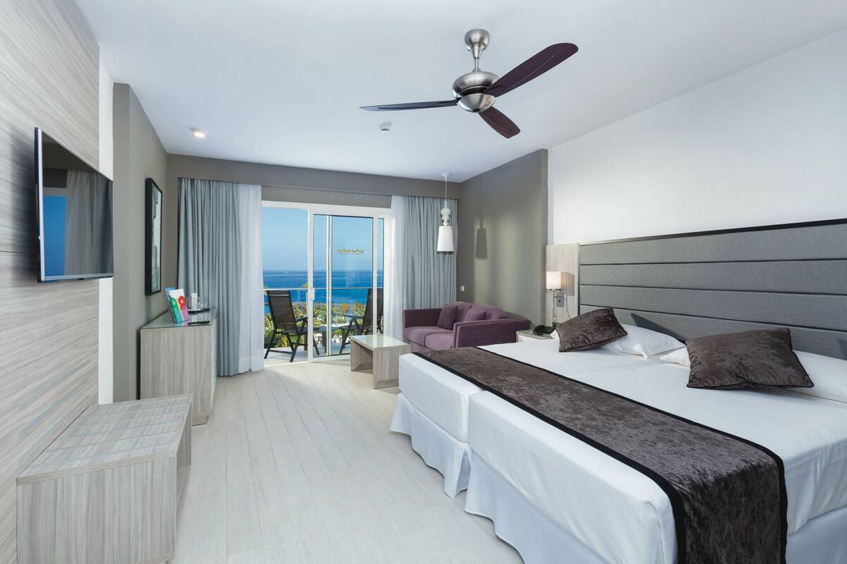 A modern hotel room with a large bed, a ceiling fan, a TV, a balcony with a view of the sea, and a seating area with a purple sofa.