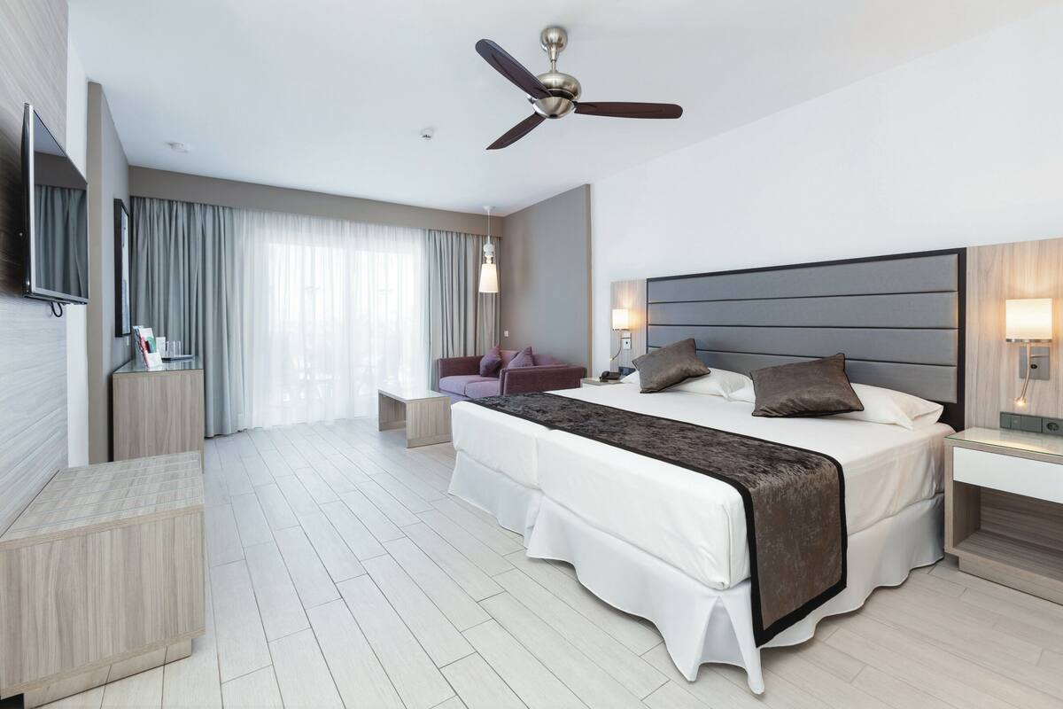 A modern hotel room with a large bed, a ceiling fan, a TV, and a desk with a lamp.
