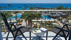 A balcony with two lounge chairs, a table with a drink and a menu, overlooking a pool area with palm trees and a view of the ocean. - Hotel Riu Palace Tenerife (Adeje)