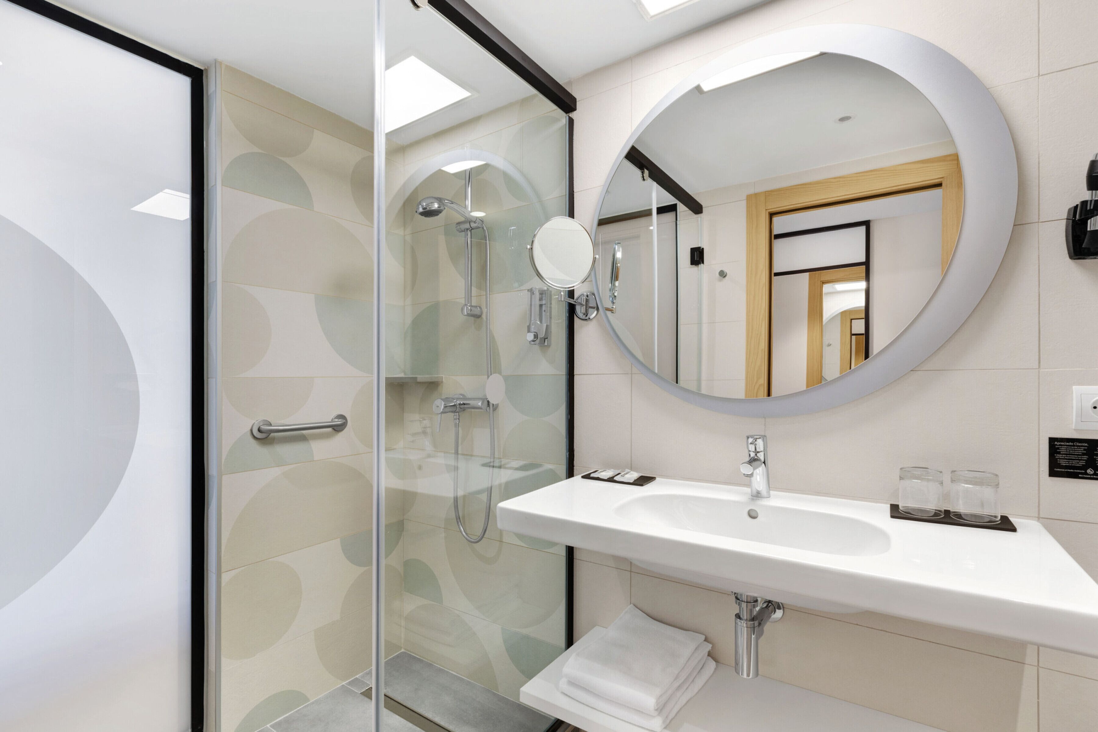 Un baño moderno con cabina de ducha de vidrio, un espejo redondo, un lavamanos blanco y un perchero para toallas.