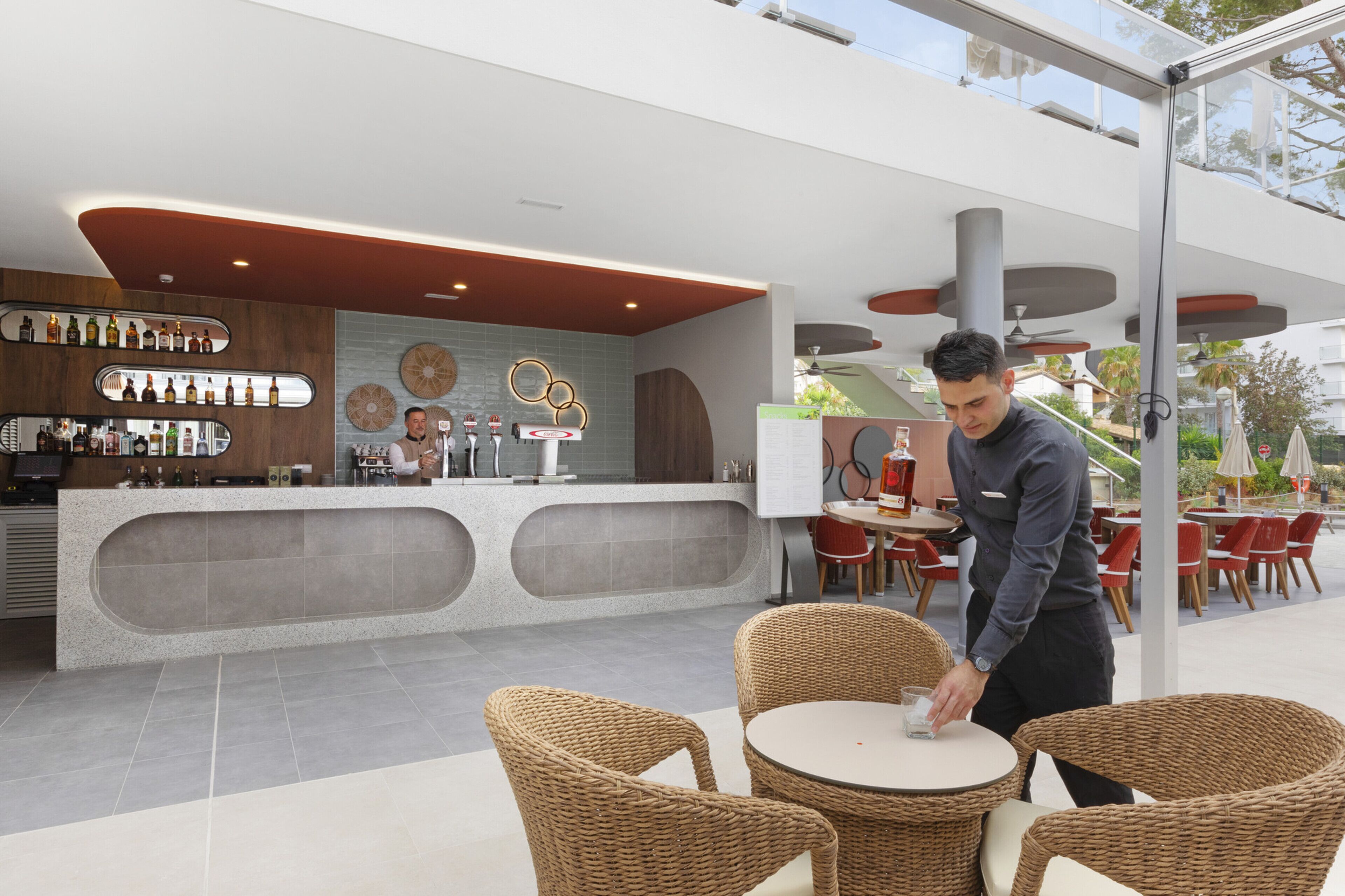 Un bar moderno con barra, taburetes y una zona de estar con sillas de mimbre.
