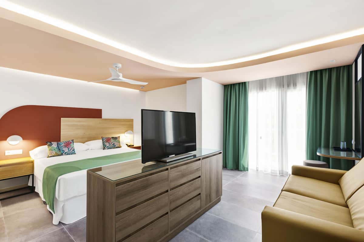 Quarto de hotel moderno com cama, televisão de tela plana, cómoda de madeira, sofá e janela grande com cortinas.