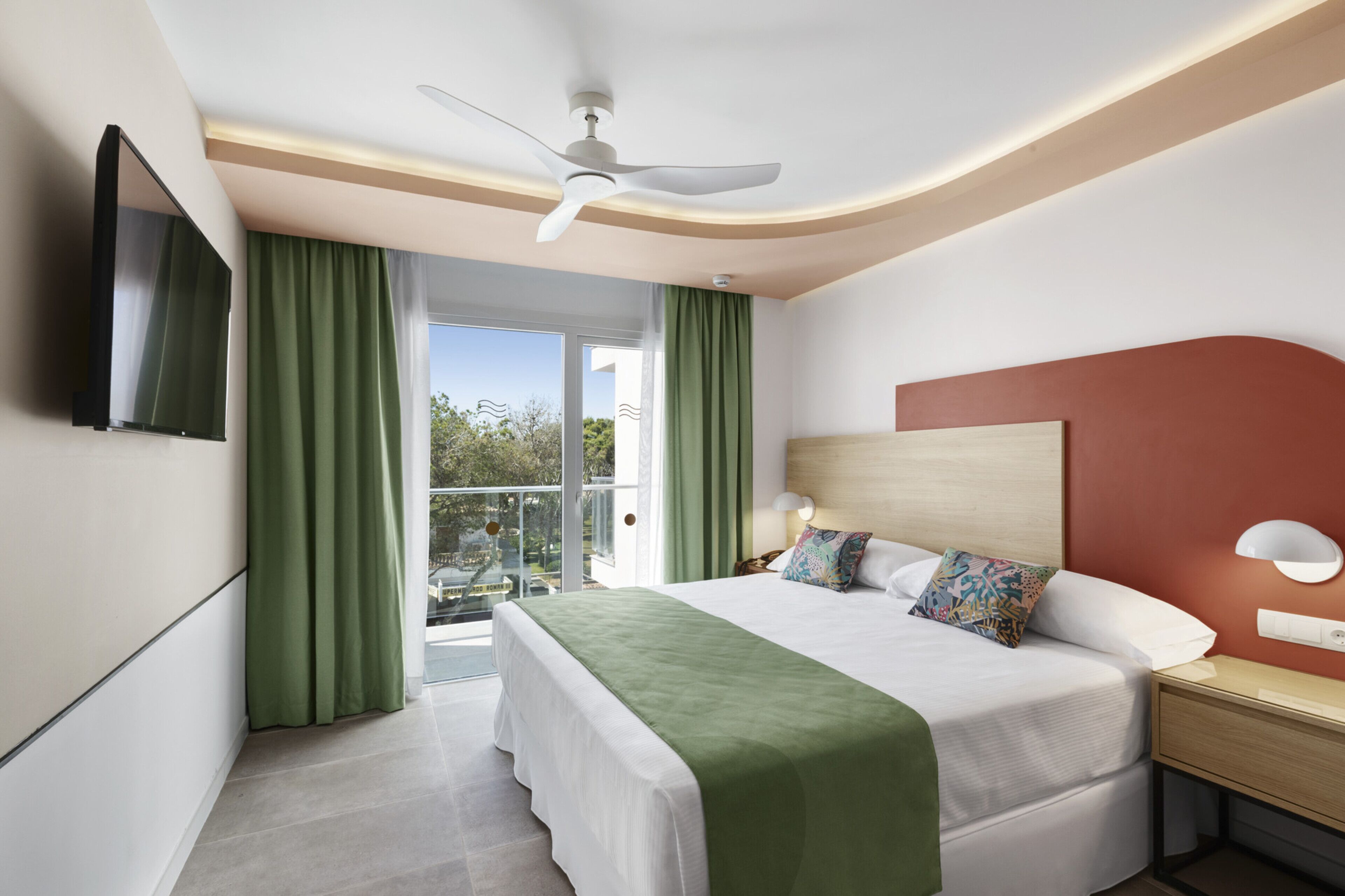 Una habitación de hotel con cama, mesita de noche, televisor y balcón con cortinas.