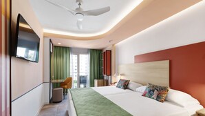 Habitación de hotel con una cama grande, un televisor de pantalla plana en la pared, un ventilador de techo, un escritorio con una silla y vistas al exterior a través de una ventana con cortinas verdes.