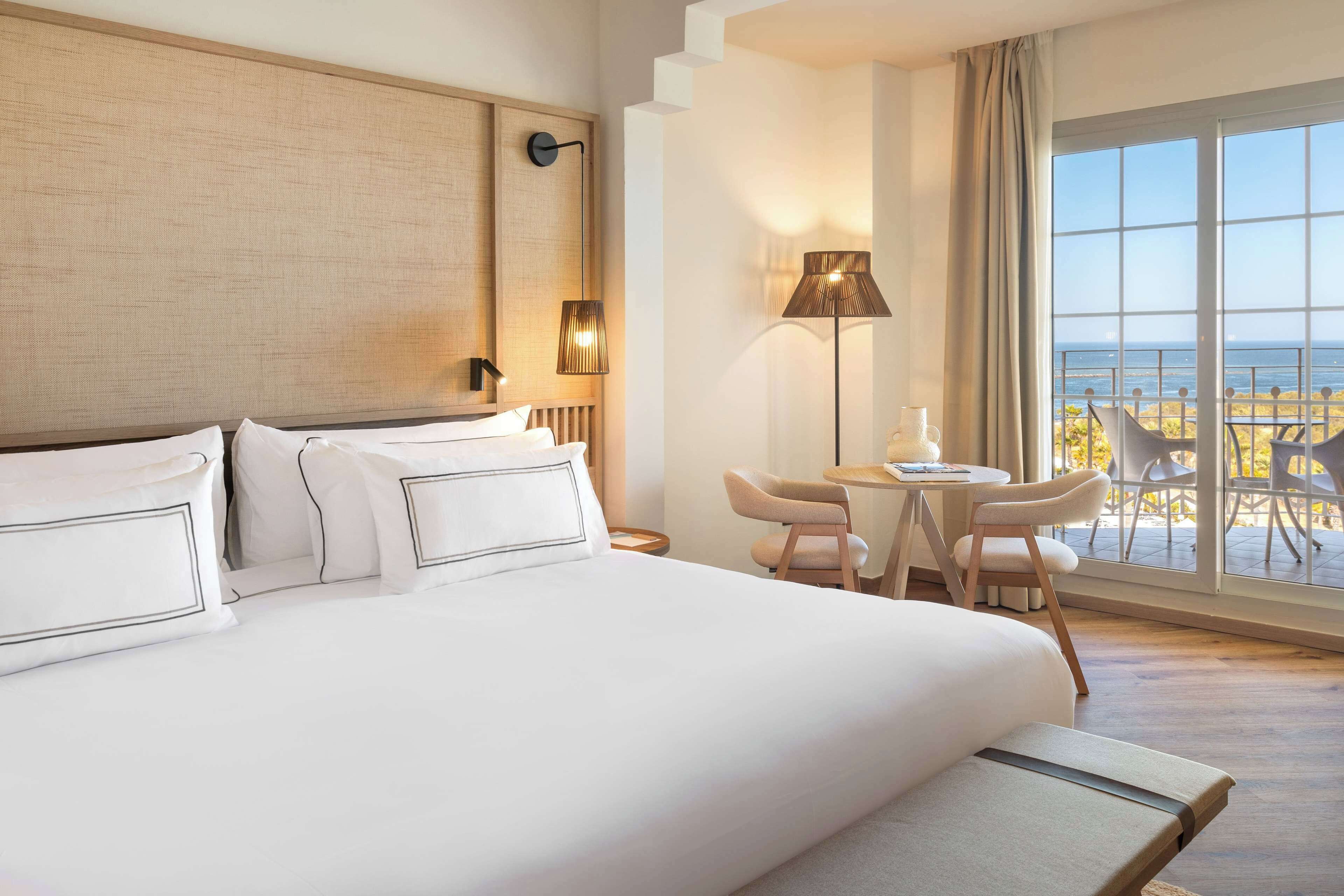 Ein Hotelzimmer mit einem großen Bett, einem Schreibtisch und Stühlen an einem großen Fenster mit Meerblick.
