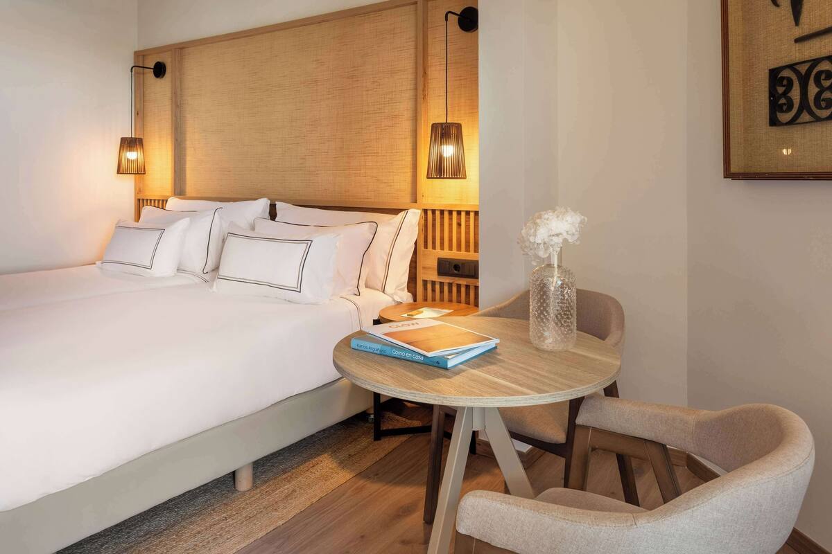 Une chambre d’hôtel comprenant un lit, des lampes de chevet, une table ronde en bois avec une chaise et un vase décoratif.