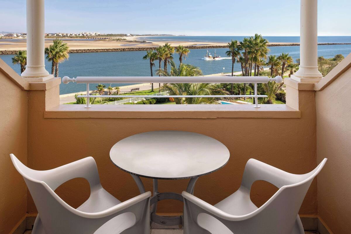 Un balcon avec une table blanche et deux chaises, donnant sur une plage bordée de palmiers et où se trouve un bateau.