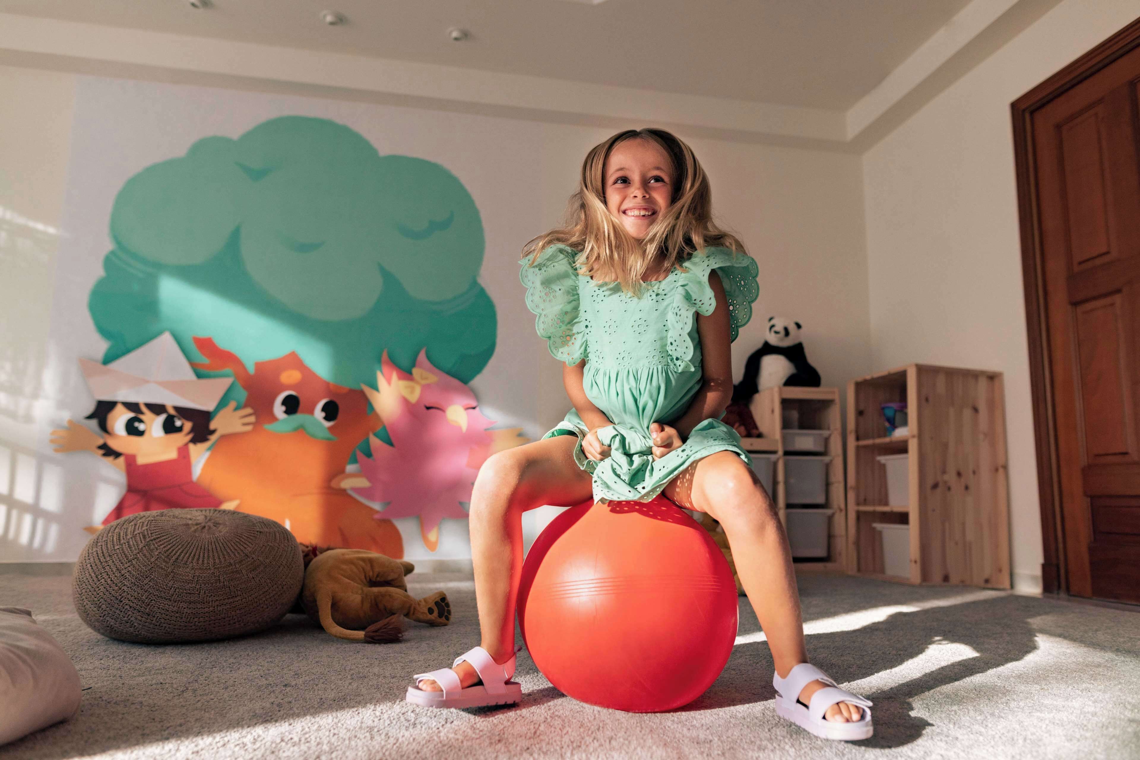 A child sitting on a red exercise ball in a room with a large cartoon mural on the wall, a wooden cabinet, and a plush toy on the floor.