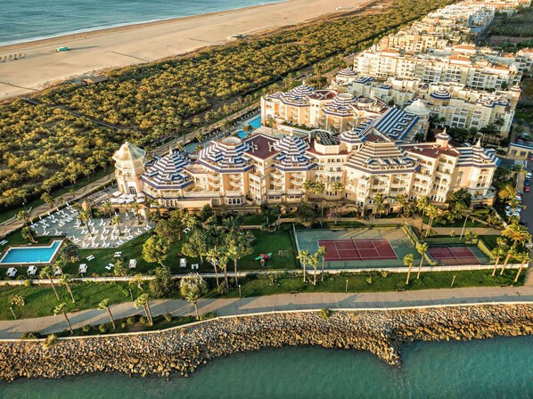 Vista aérea de un complejo turístico con varias edificaciones, piscina, canchas de tenis y una playa.