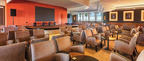 Un salón de conferencias con filas de sillas, un escenario con una pantalla grande y una zona de estar con mesas.