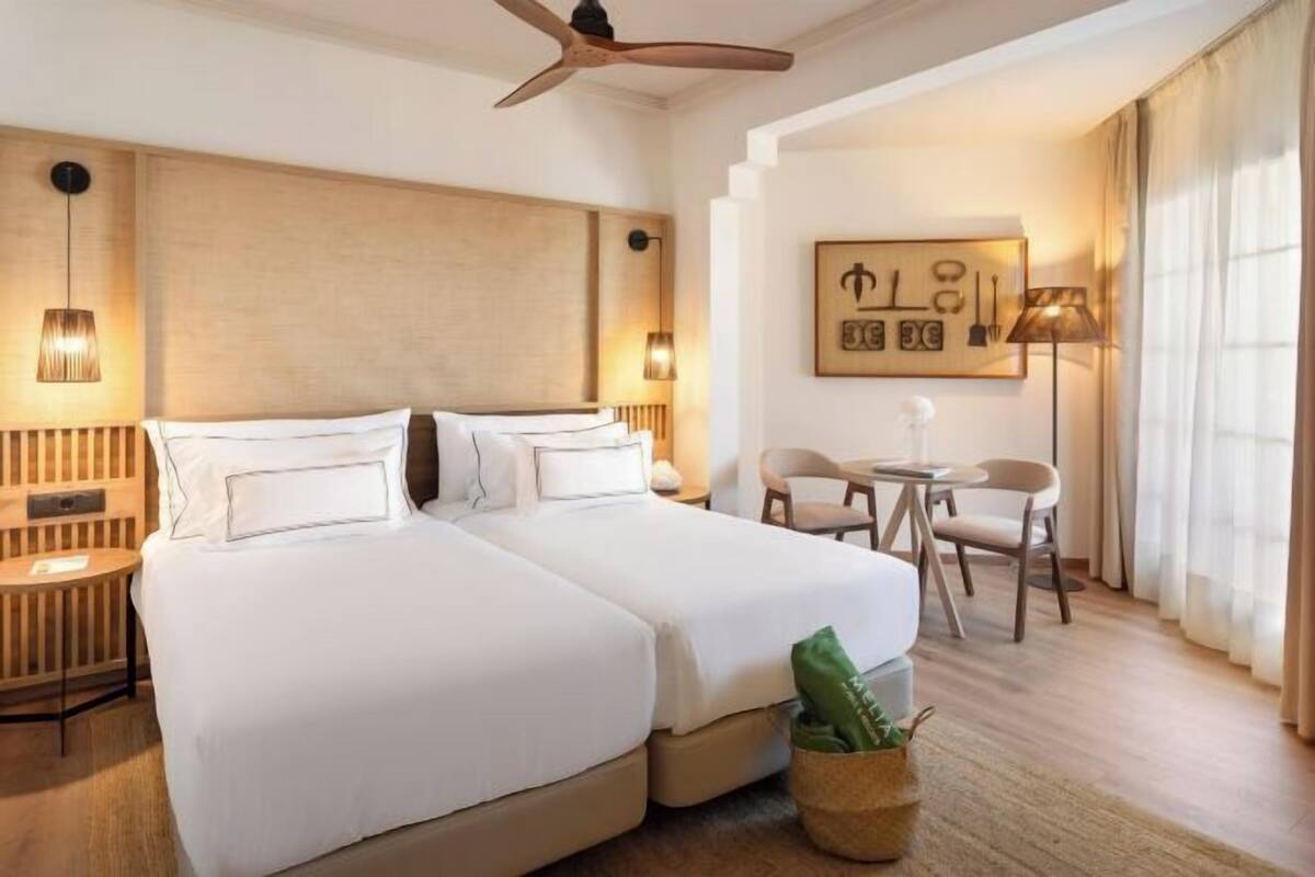 Une chambre d’hôtel avec un grand lit, un bureau, une chaise, un ventilateur de plafond et une lampe fixée au mur.