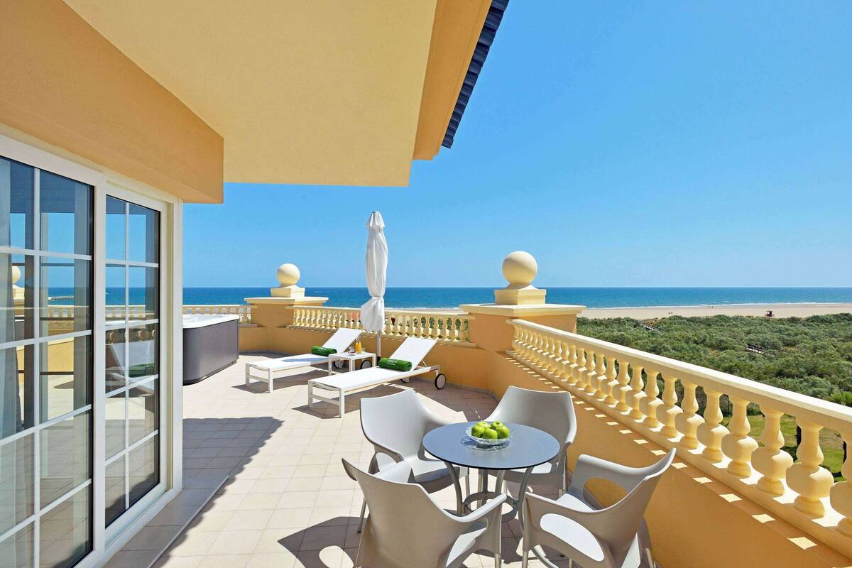 Un balcon avec vue sur la plage, des chaises blanches, une table avec des fruits et un parasol.