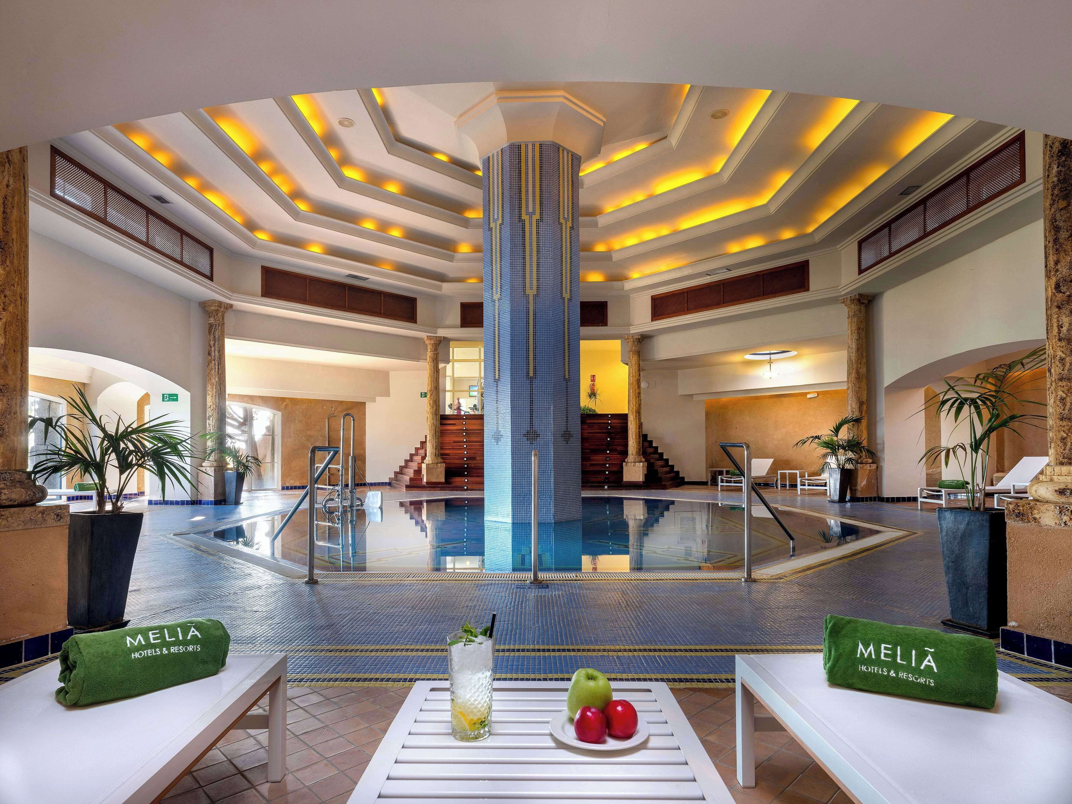 Vestíbulo de hotel con piscina central, una escalera y zonas de estar con toallas y fruta.