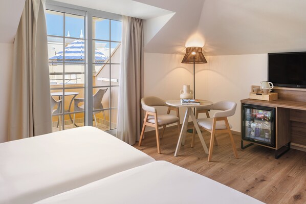 A hotel room with a bed, a desk with a TV, a refrigerator, a lamp, and a view of a building with a blue roof.