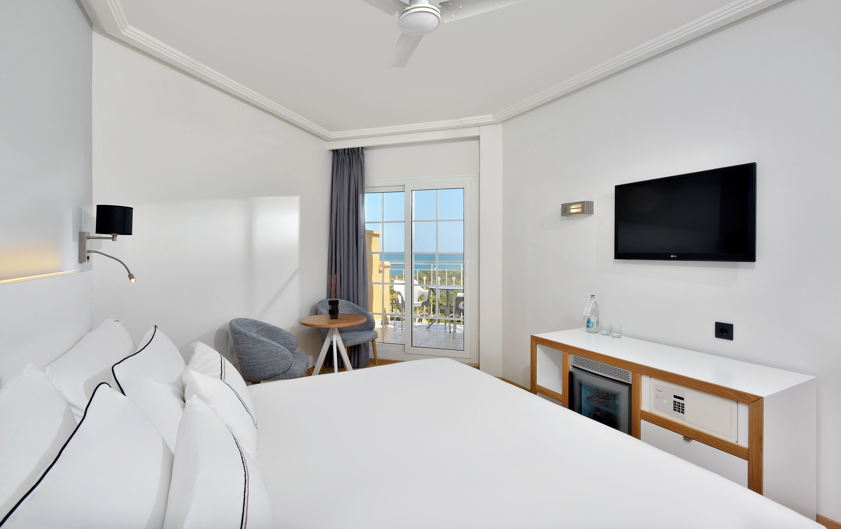 Habitación de hotel con cama, escritorio con televisor, una pequeña mesa con sillas y vista al mar.