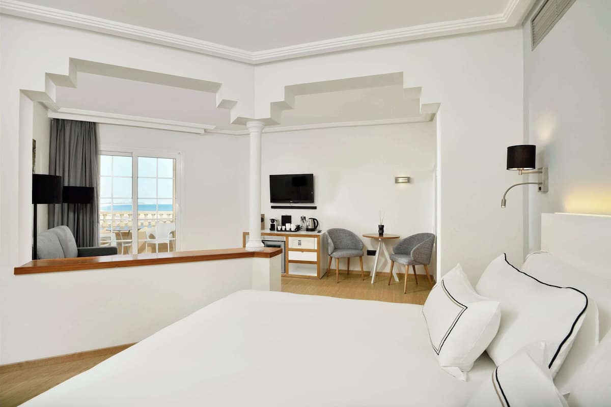 Une chambre d’hôtel moderne avec un grand lit, un bureau avec une télévision et une vue sur la plage par la fenêtre.