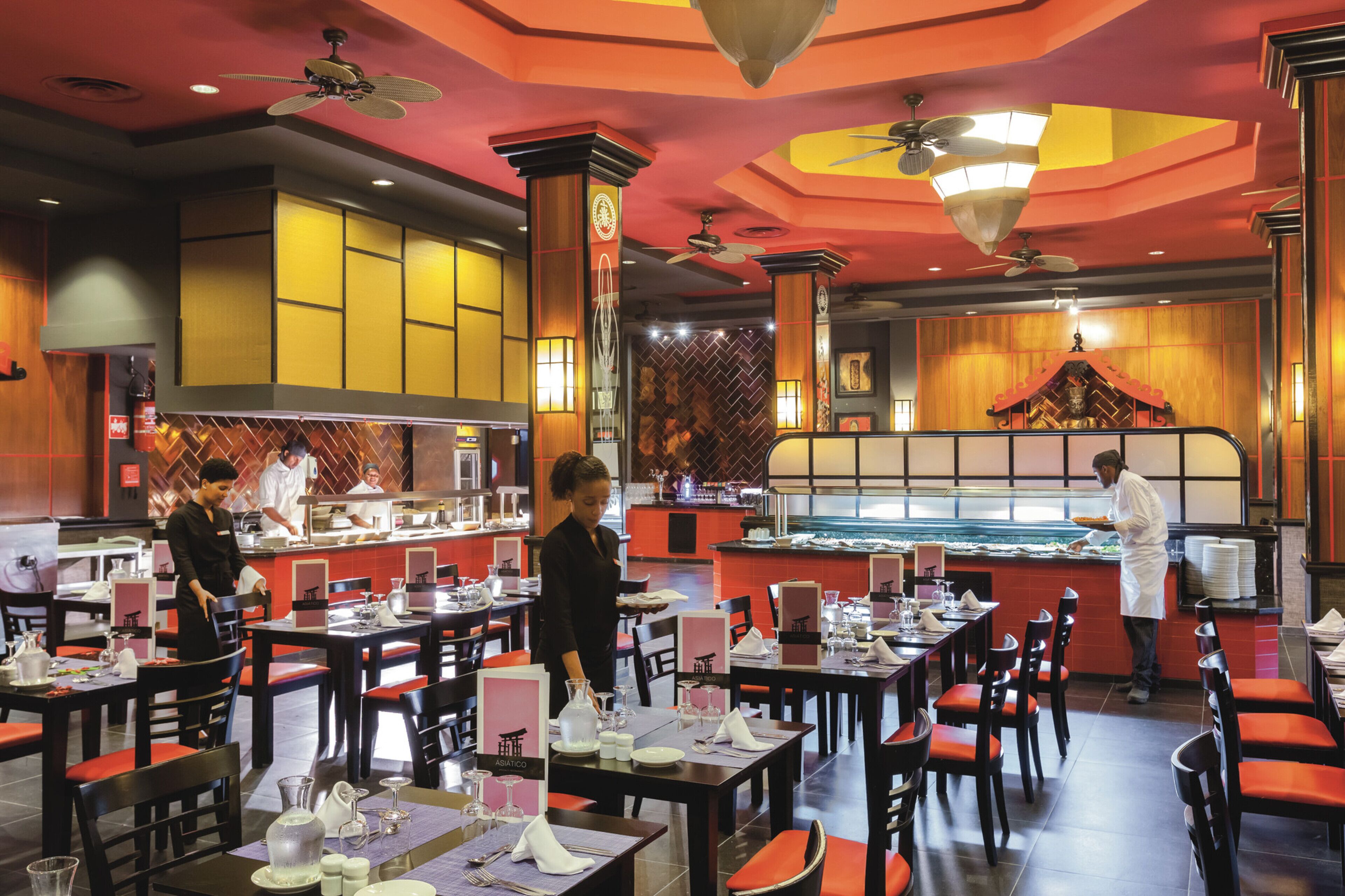 A restaurant with multiple tables and chairs, a counter with a chef, and a decorative ceiling.
