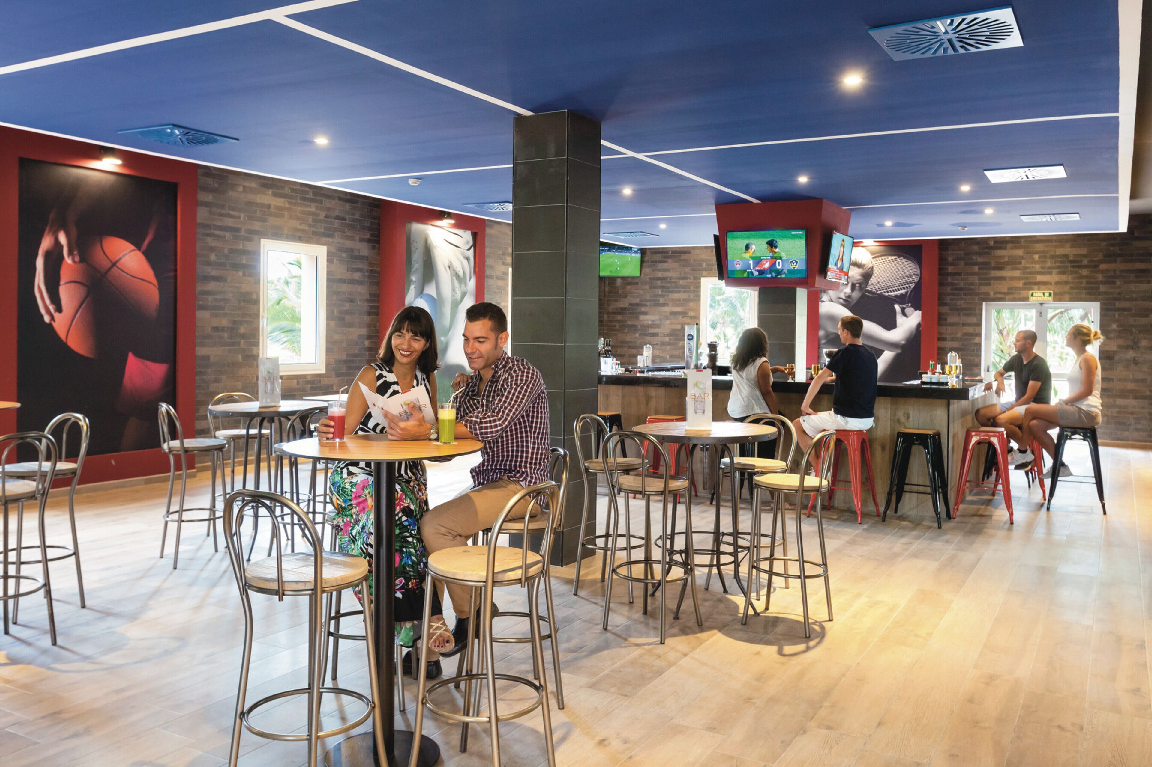 Un área de comedor interior moderna con varias mesas y sillas, una barra con estación de bebidas y pantallas grandes que muestran eventos deportivos.