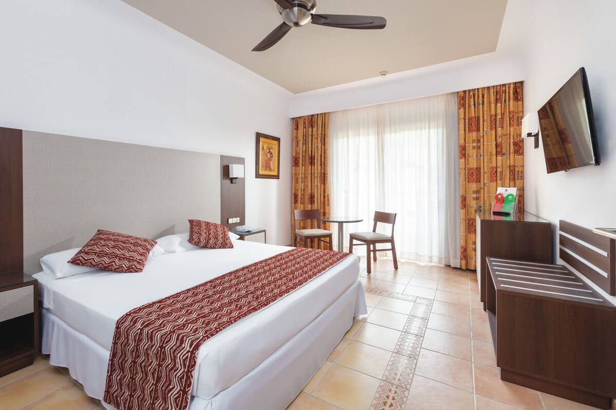 A hotel room with a large bed, a desk, a chair, a TV, and a ceiling fan.