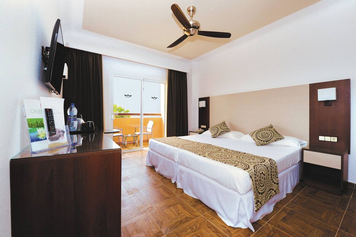 A hotel room with a bed, a desk with a computer, a ceiling fan, and a view of the outdoors.