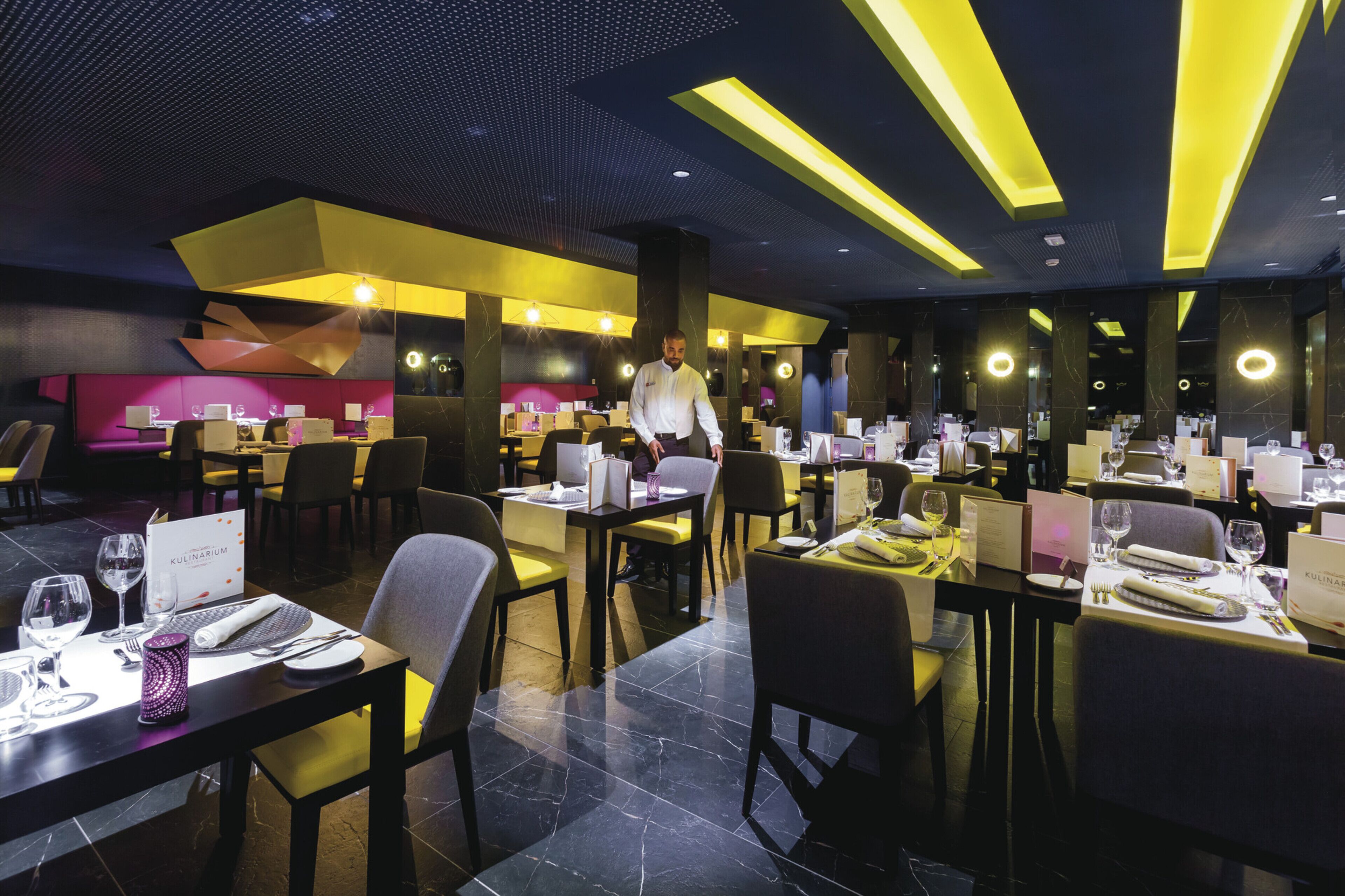 A modern restaurant interior with multiple dining tables, chairs, and a waitstaff member.