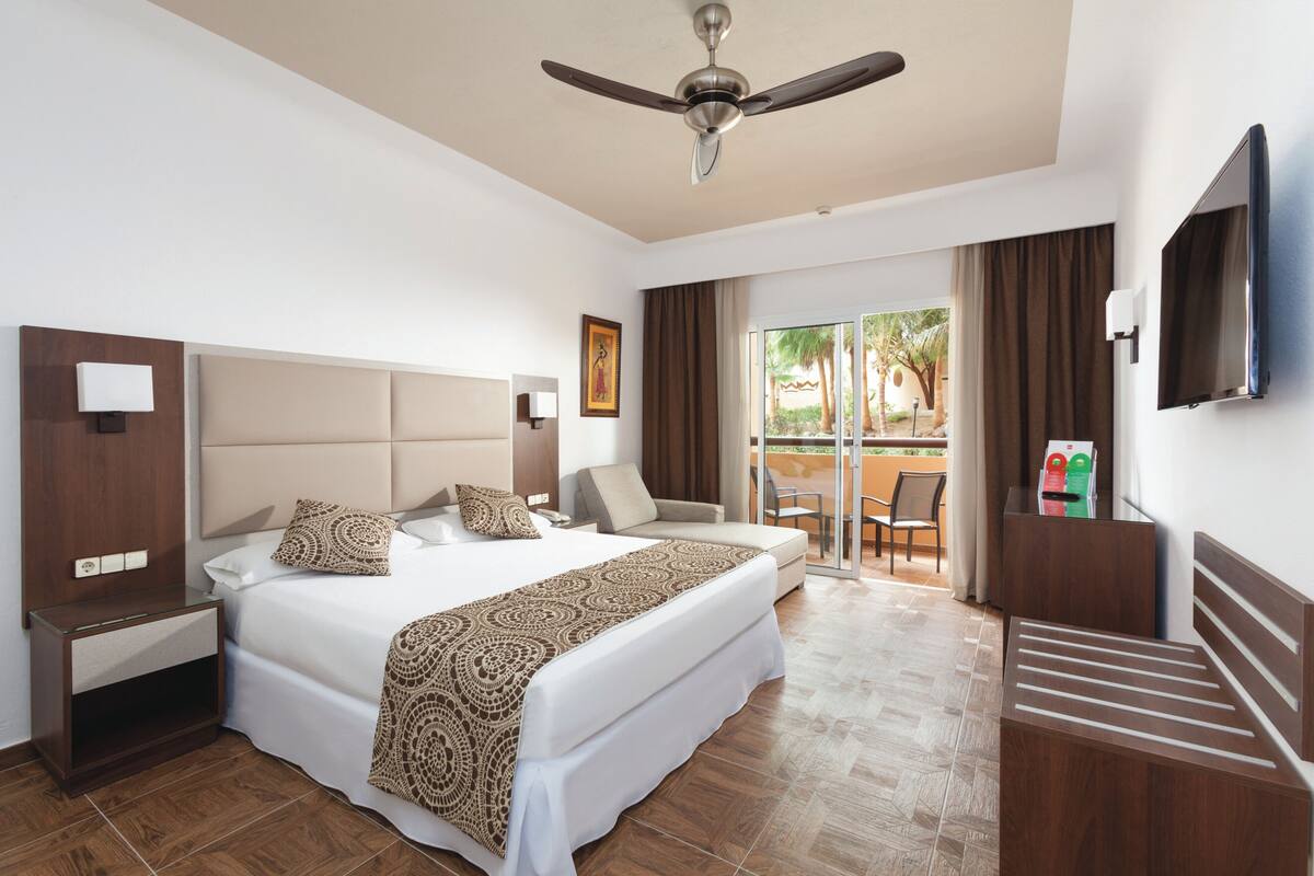 A hotel room with a large bed, a ceiling fan, a TV, a desk, and a view of the outside.