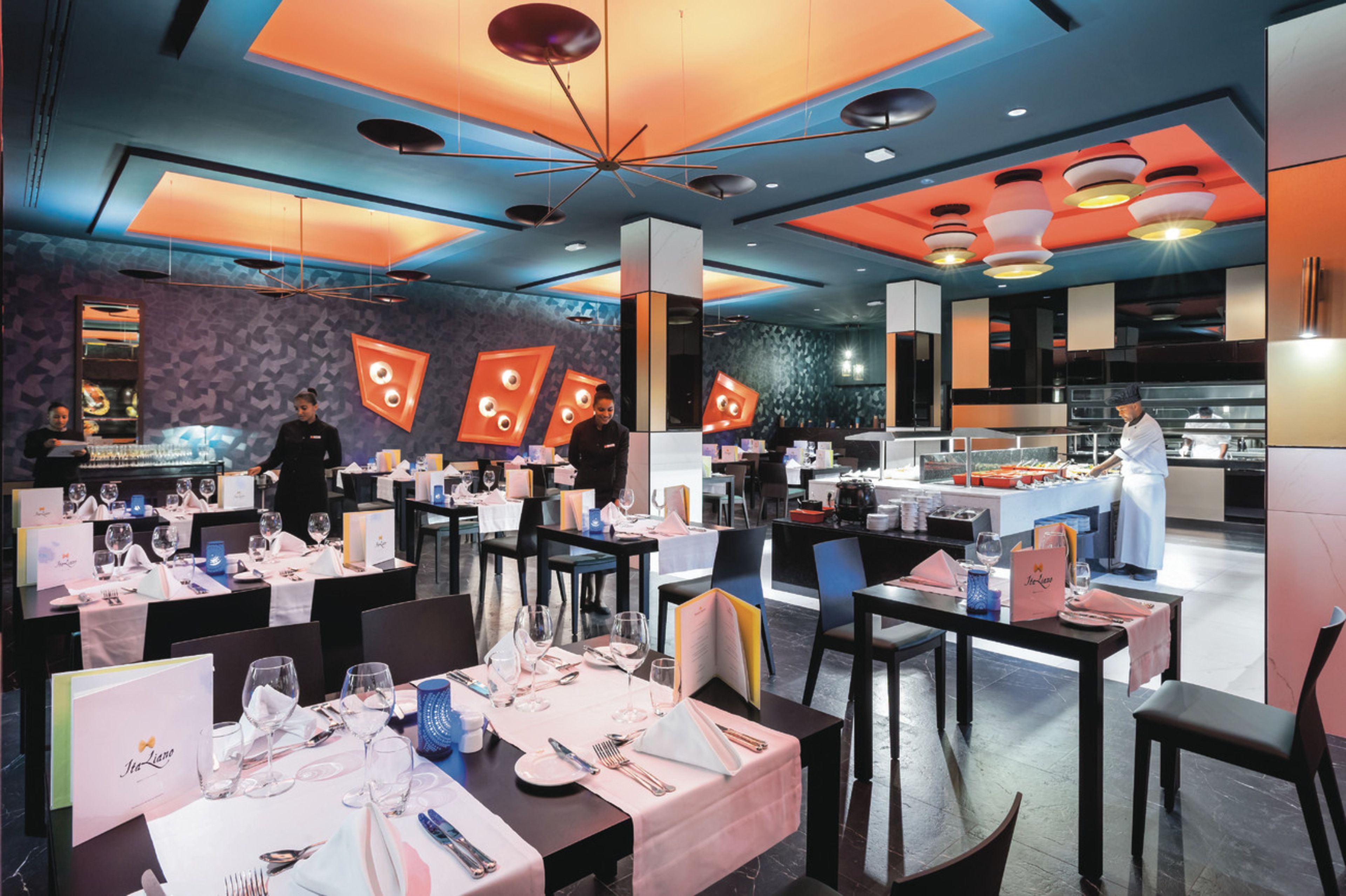Un interior de restaurante moderno con varias mesas, sillas y una zona de cocina al fondo.