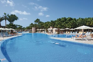 A large swimming pool with lounge chairs and umbrellas, surrounded by palm trees.