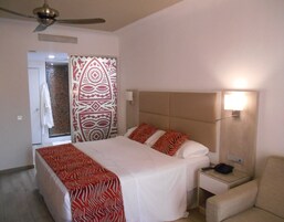 Habitación de hotel con cama, mesita de noche, una silla, ventilador de techo y zona de ducha con cortina estampada.