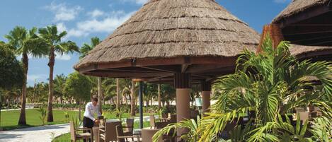 A covered outdoor seating area with thatched umbrellas, wicker furniture, and palm trees.