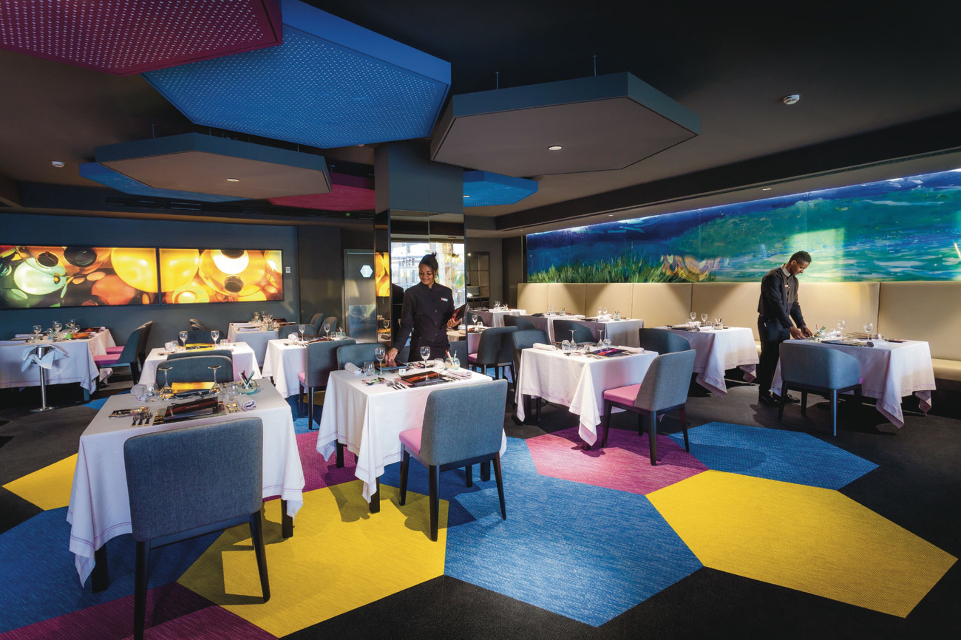 A modern restaurant with multiple tables, a large aquarium wall, and a colorful floor design.