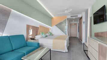 Een moderne hotelkamer met een bank, een bed met een beige deken, een aan de muur bevestigde televisie en een plafondventilator.