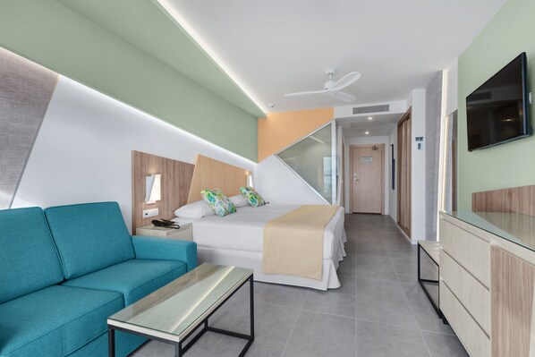 A modern hotel room with a sofa, a bed with a beige blanket, a TV mounted on the wall, and a ceiling fan.