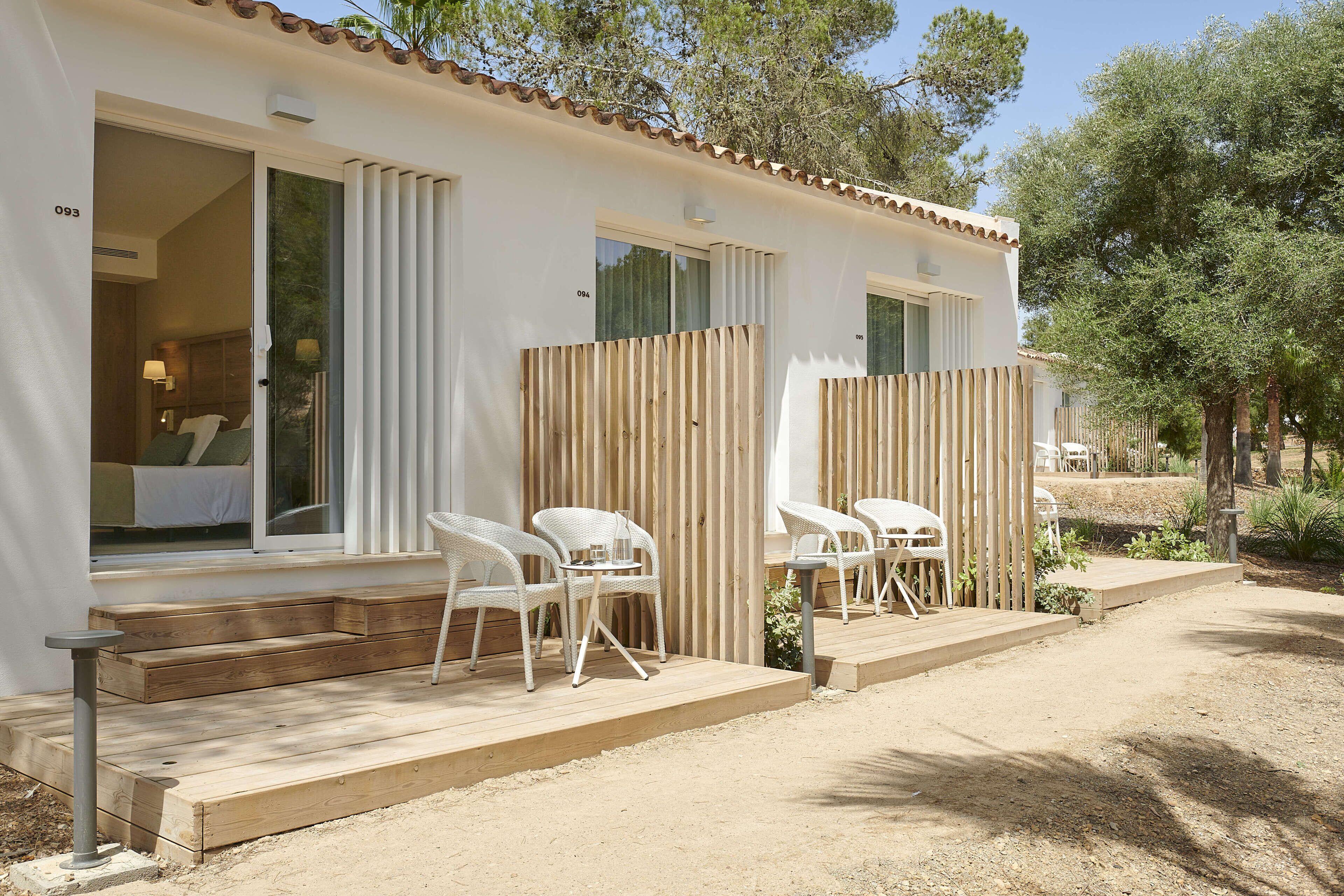 Una villa blanca con una terraza de madera, dos sillas blancas de mimbre y una pequeña mesa.