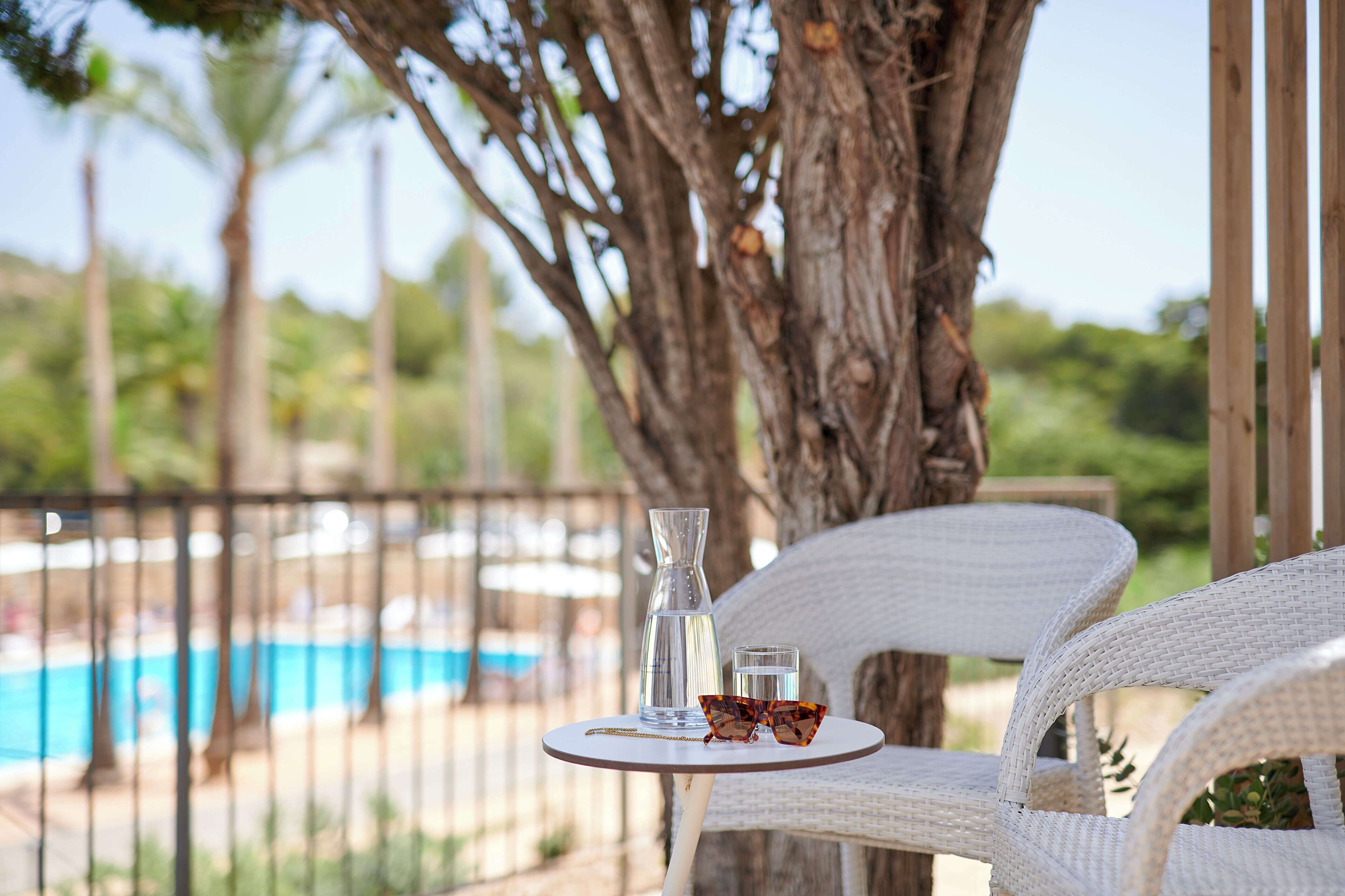 Silla de mimbre y mesa con un vaso y gafas de sol, con vista a una piscina.