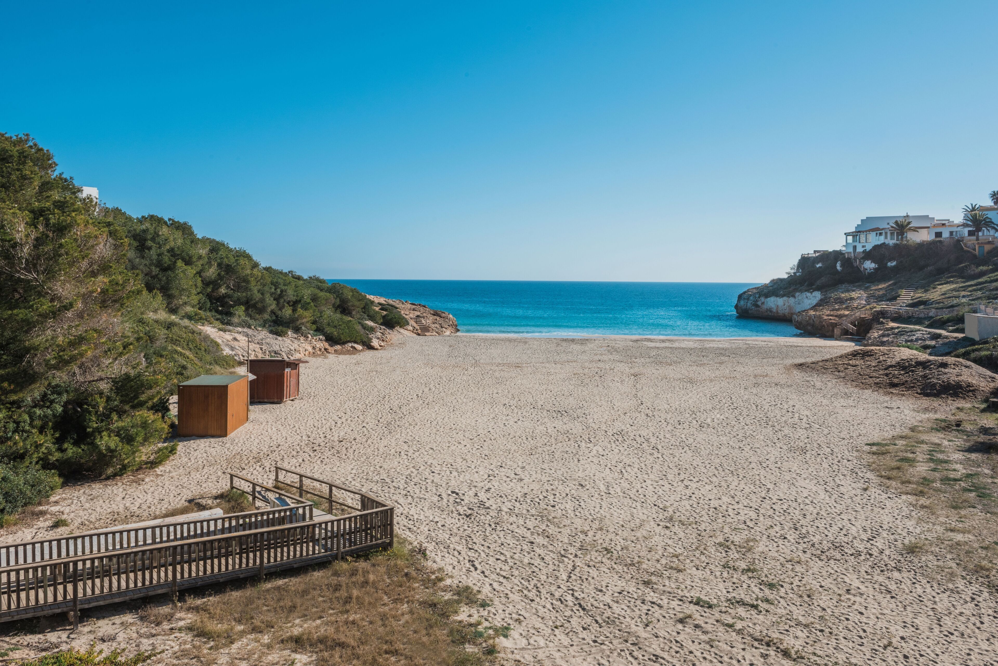 A beach with a wooden walkway leading to a wooden structure, a clear blue sky, and a building on a cliff in the background.