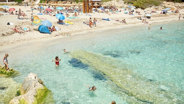 Une plage avec une eau cristalline, des gens qui se baignent et prennent des bains de soleil, et des parasols sur le sable.