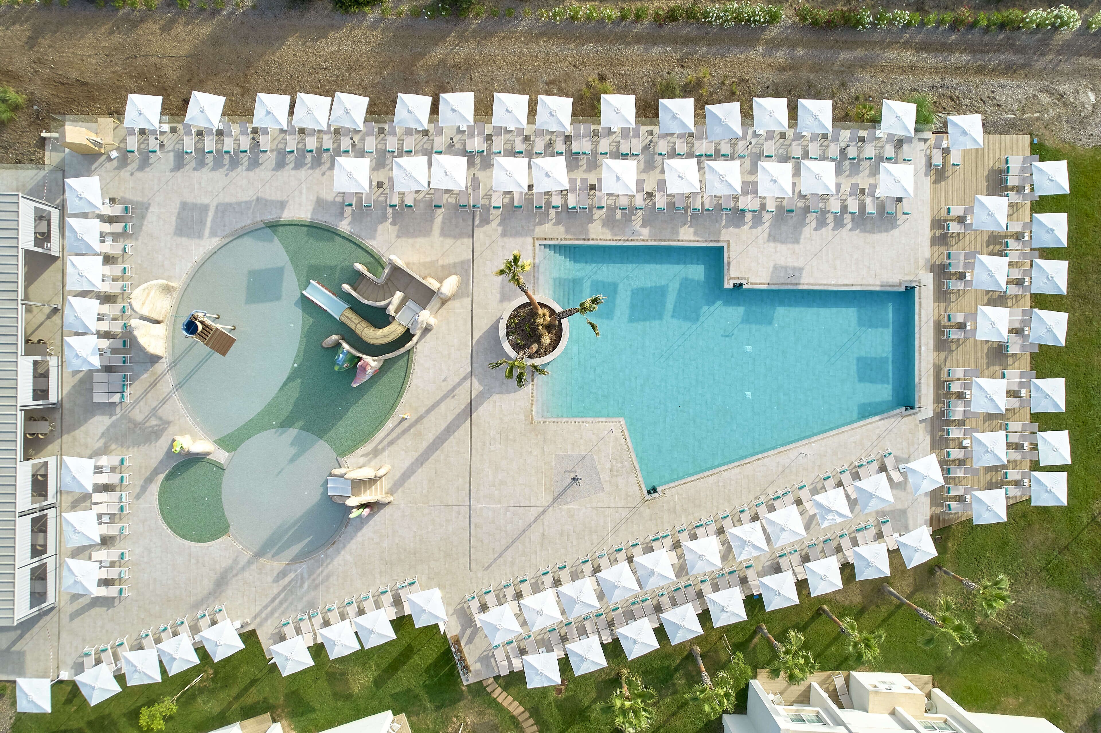 An aerial view of a resort with a large swimming pool, sun loungers, and umbrellas arranged around the pool area.