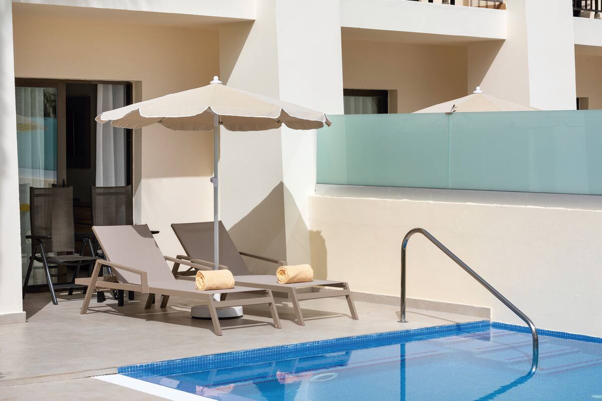 Zona de piscina amb gandules i ombrelles, una piscina amb rajoles blaves i una barana de vidre.