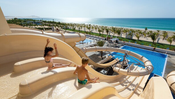 A water slide at a resort with a pool and beach view. - Hotel Riu Karamboa - Adults Only - All Inclusive (Boa Vista)
