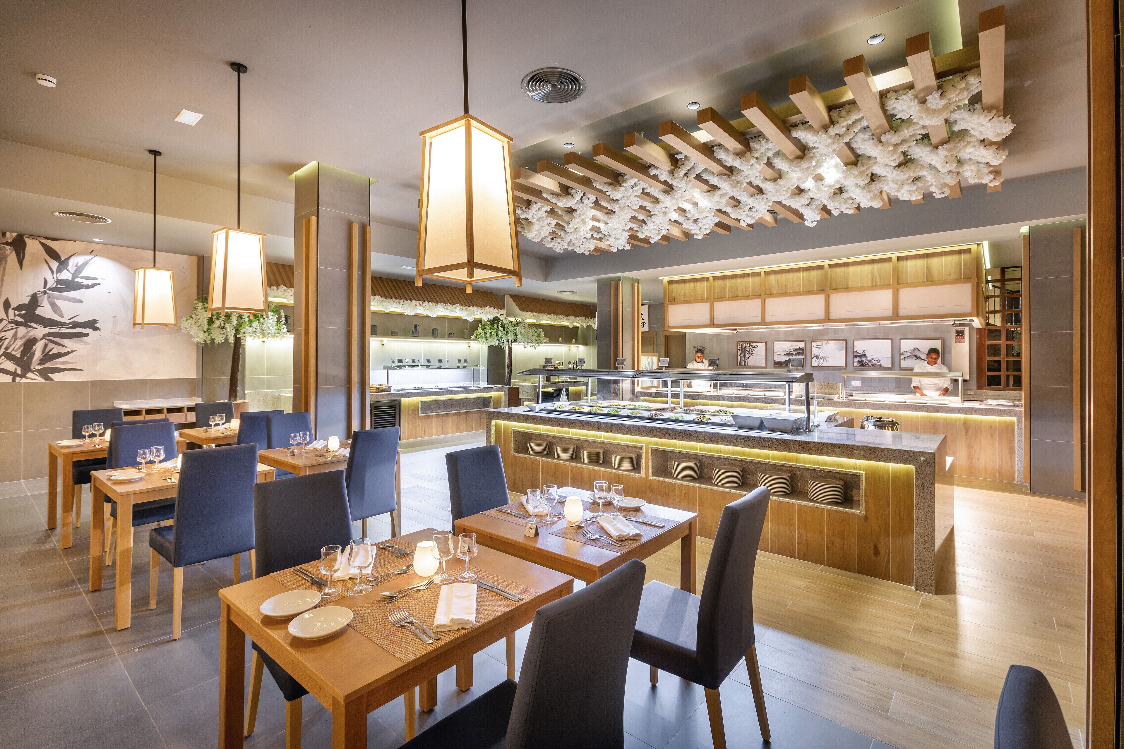 Ein modernes Restaurant mit Holztischen und -stühlen, einer zentralen Kochinsel mit Theke und hängenden Pendelleuchten.