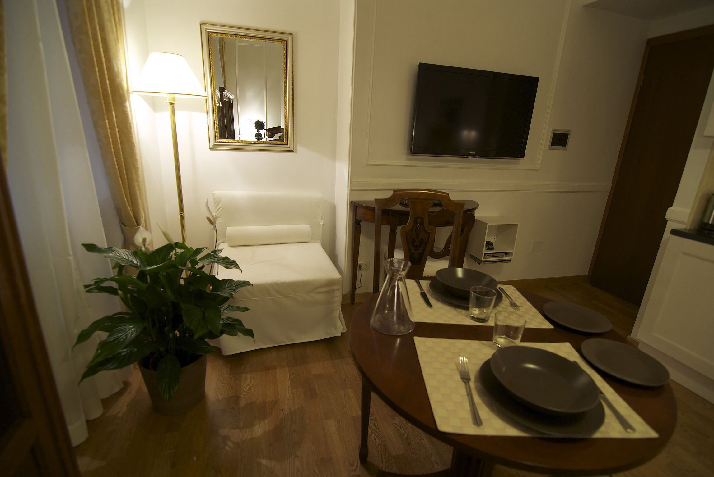 A hotel room with a bed, a dining table set with plates, a chair, a potted plant, a lamp, and a TV mounted on the wall.