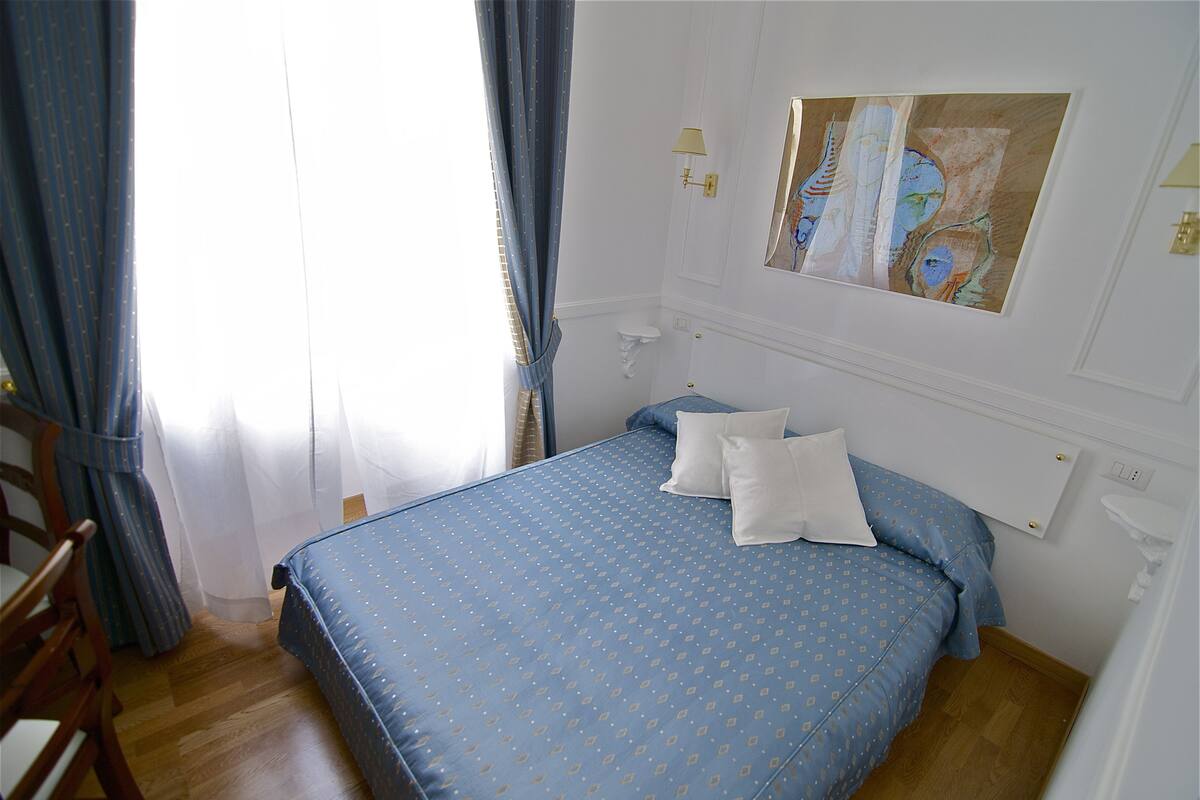 Une chambre à coucher avec un lit, deux oreillers, une chaise en bois, un mur blanc, un tableau et une fenêtre avec des rideaux.