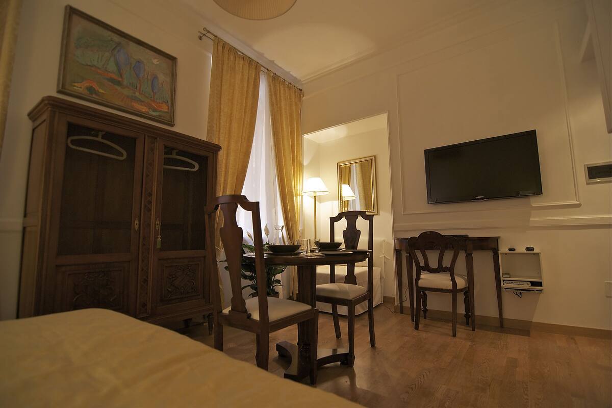Une chambre d’hôtel comprenant un lit, une armoire en bois, une table à manger avec des chaises, un téléviseur fixé au mur et une fenêtre avec des rideaux.