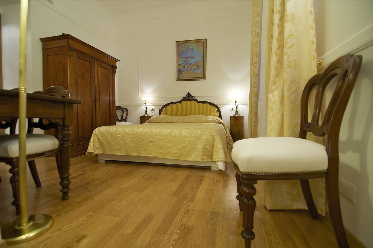 Une chambre avec un plancher en bois, un lit recouvert d’une couverture jaune, une chaise en bois, une armoire en bois et un tableau accroché au mur.