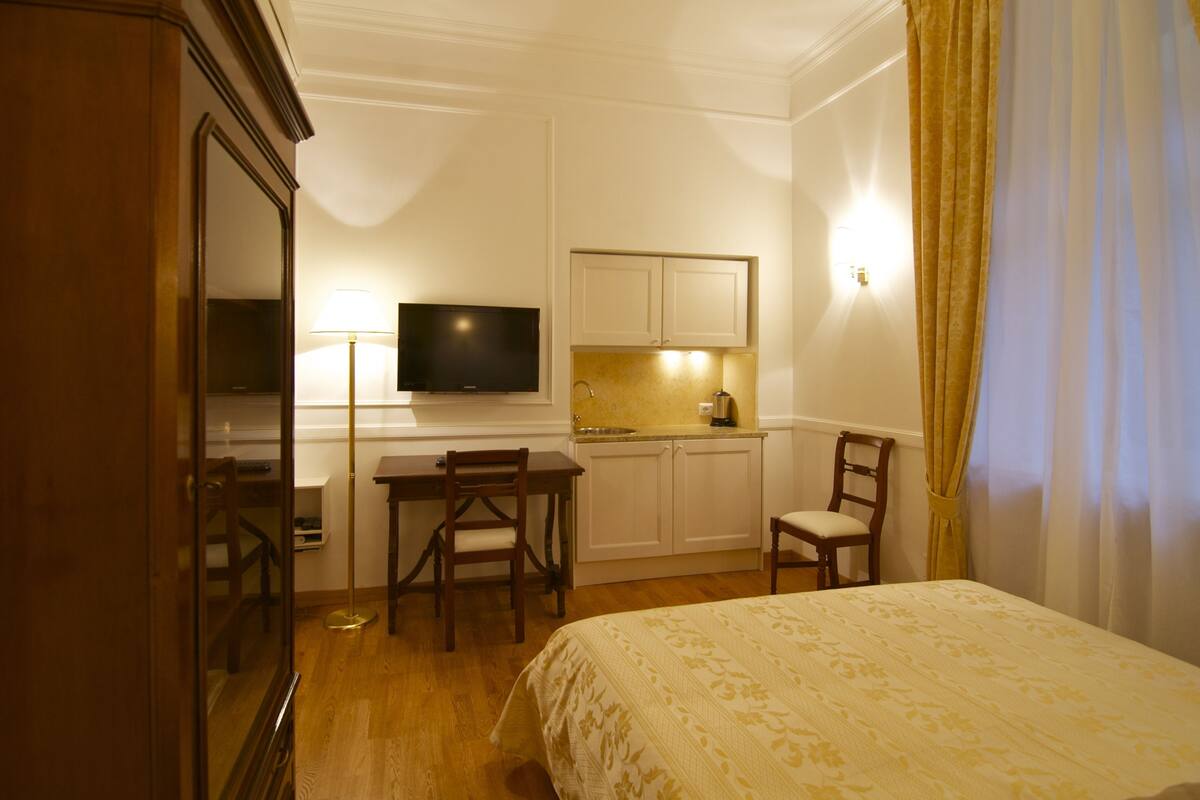 Une chambre d’hôtel comprenant un lit, un bureau avec une chaise, une télévision, une petite kitchenette et un miroir.