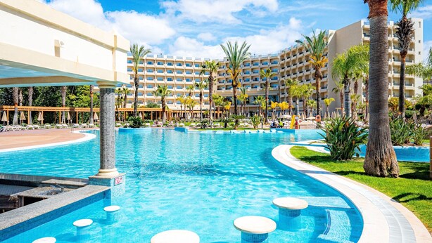 A hotel pool area with a large pool, palm trees, and a multi-story building in the background. - 3HB Guarana (Albufeira)