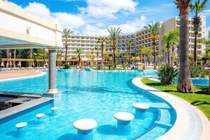A hotel pool area with a large pool, palm trees, and a multi-story building in the background.